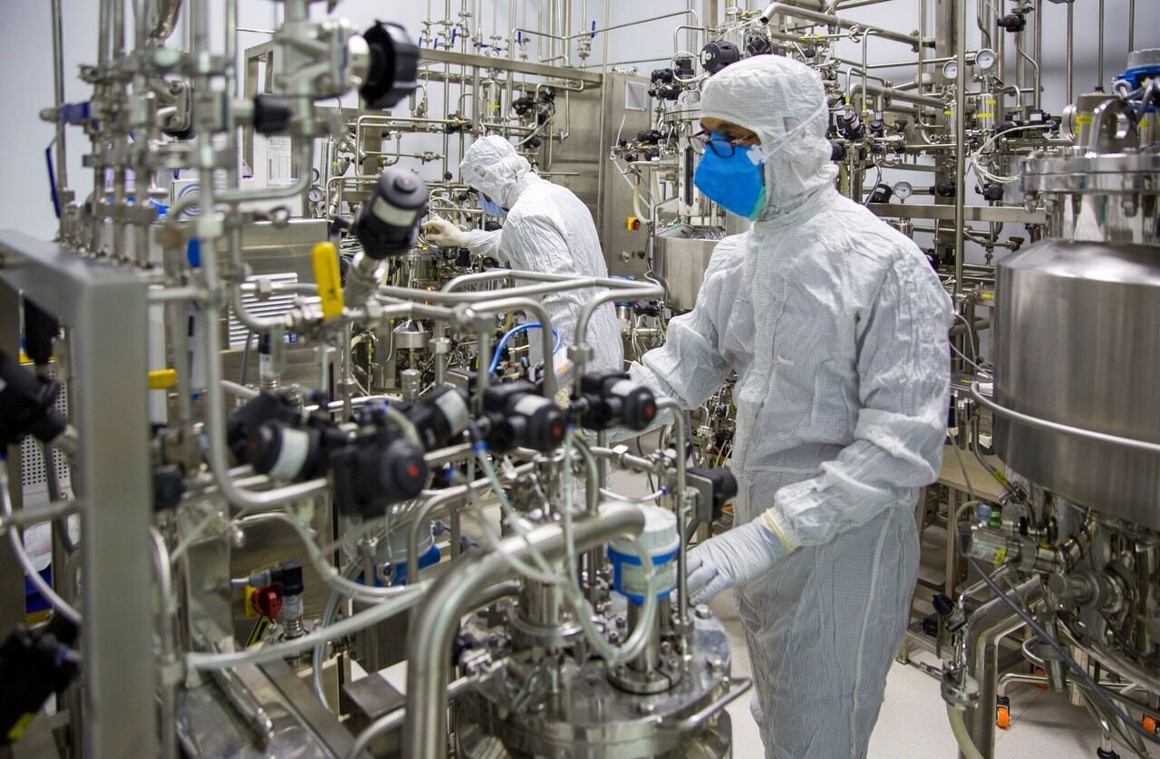 Two operators in white cleanroom suits, hoods, and masks work among stainless-steel bioreactors and dense process piping inside a biopharmaceutical production suite.