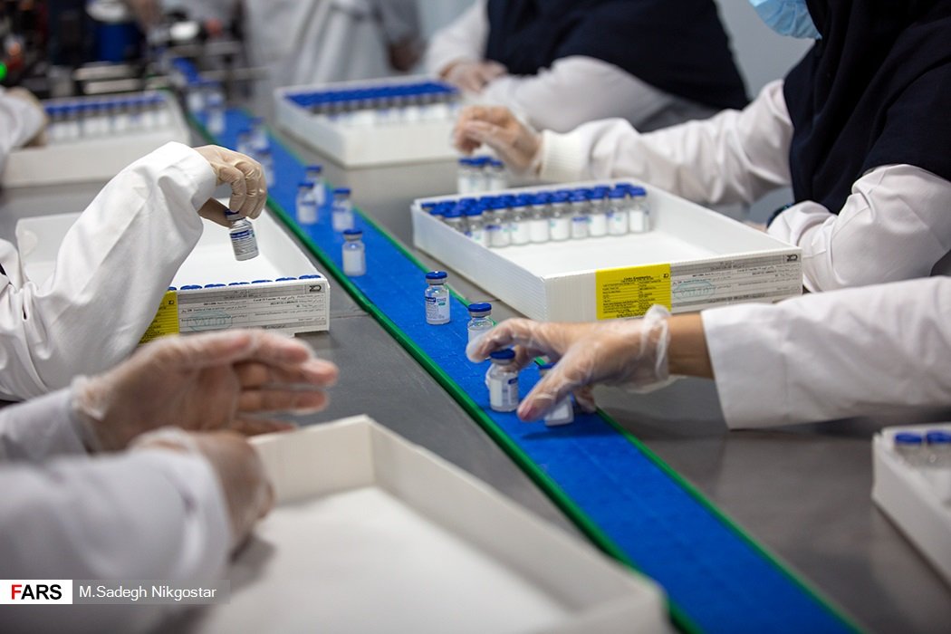 Gloved workers pick labeled vials off a conveyor belt and pack them into printed cartons on a pharmaceutical packaging line.