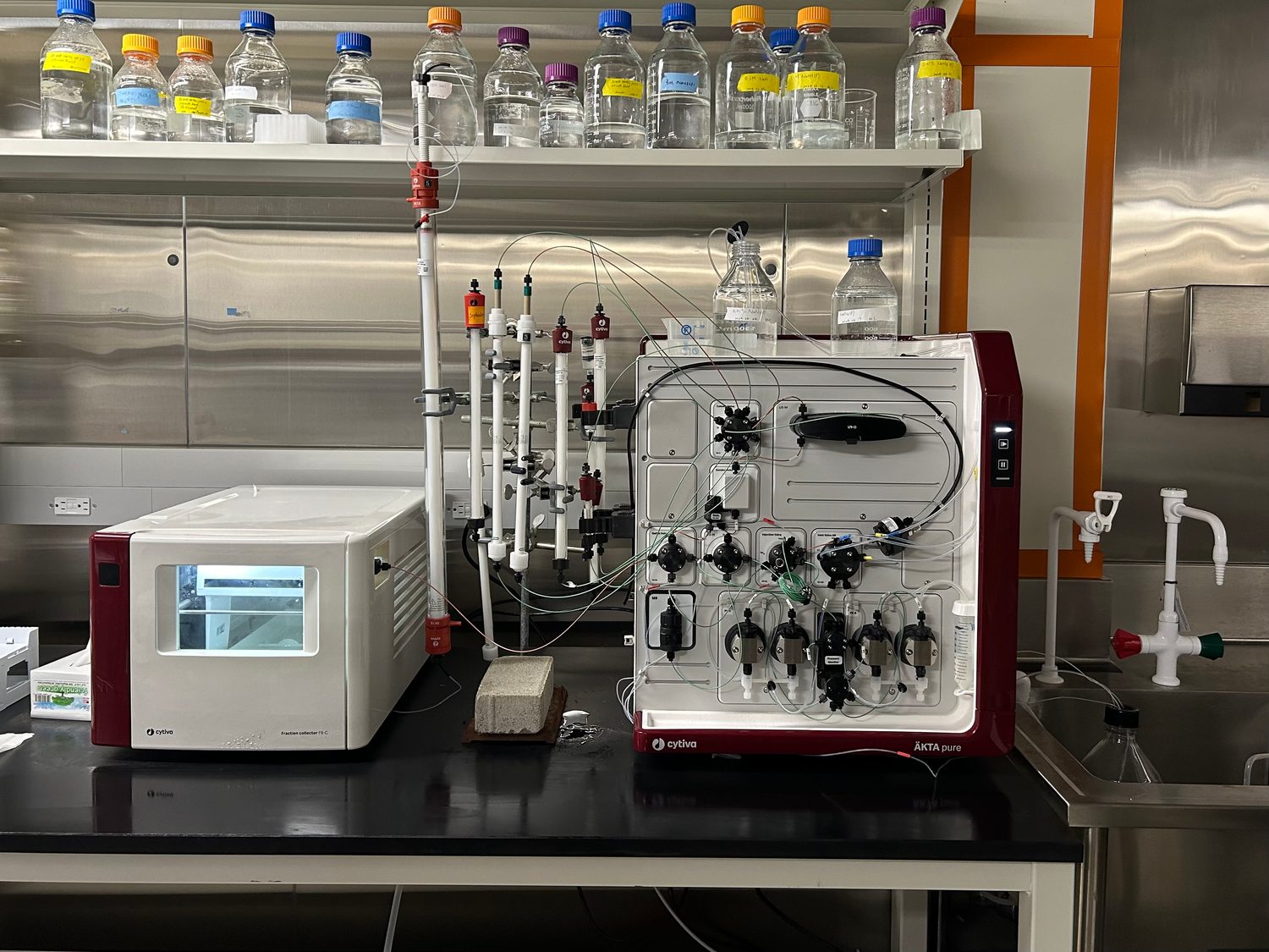 A benchtop ÄKTA pure FPLC chromatography system in a maroon-and-white housing beside several clear chromatography columns clamped upright on a stand, with buffer bottles on a shelf above.